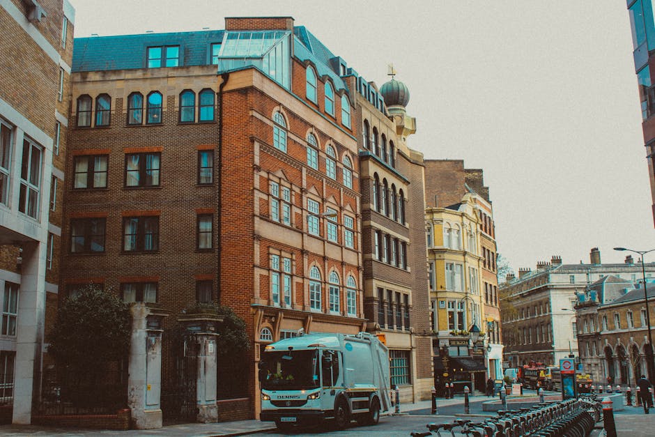A city street scene showing a row of multi-story buildings with diverse architectural styles, including a prominent red-brick building with arched windows and a decorative facade, alongside lighter-colored buildings with ornate detailing and bay windows. On the foreground, a small waste collection truck with a metallic silver finish, orange hazard lights, and branding identifiable as Waste Removal St John's Wood is parked near the curb, ready for a rubbish collection or clearance. The street surface is paved, with a designated bike lane visible on the right side, featuring a series of bike racks. Pedestrian bollards and street lamps line the walkway, and in the background, more historic buildings and modern structures create an urban environment typical of central London. The lighting appears natural with an overcast sky, emphasizing a professional and neutral tone appropriate to a waste removal service context, subtly connecting to private rubbish collection activities in the area.