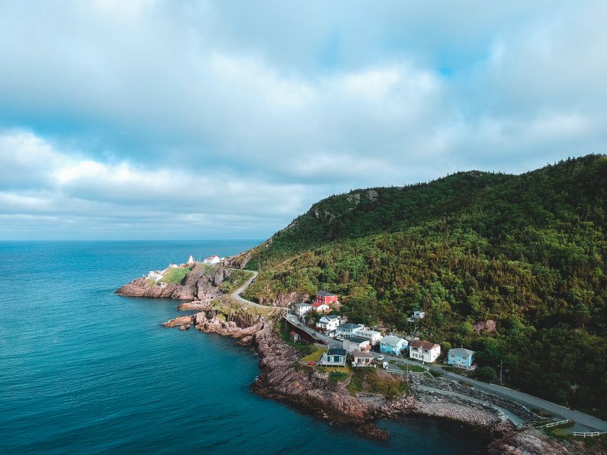 An aerial view of a coastal hillside showing a winding road with small, colorful residential houses positioned along the shoreline below a lush, densely wooded hill. The buildings are primarily white, with some featuring blue and red facades, and have either flat or pitched roofs with various finishes such as tiles or shingles. The rocky coastline is visible, with jagged rocks extending into the calm, turquoise sea, while the sky overhead is partly cloudy with patches of blue. The environment suggests a serene seaside village setting, typical of rural or semi-urban coastal areas. This image may be used to illustrate the contrast between natural landscapes and human habitation, subtly reinforcing themes of land and property management, relevant to private waste removal services such as those provided by Waste Removal St John's Wood on residential areas near coastlines, highlighting the importance of effective rubbish clearance in picturesque settings.