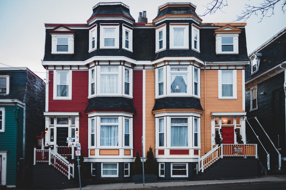 A three-storey Victorian-style residential building with a distinctive bay window design, painted in vibrant contrasting colours—red on the left side and a warm mustard yellow on the right. The façade features multiple white-framed sash windows, some with curtains visible inside, and decorative elements such as cornices and trims. The building's roof has a dark slate finish with multiple pitched sections, rounded corner features, and small dormer windows. The front entrance is accessible via a short, wide staircase with white-painted railings, leading to a black front door flanked by small ornamental shrubs. To the left of the entrance is a lamppost, and the pavement in front is made of concrete slabs, with a small lawn and a few young trees nearby. The scene is captured during daylight with a predominantly clear sky, emphasizing the building’s colorful exterior and architectural details, positioned amidst neighboring houses with similar Victorian features, underscoring an urban residential setting that may require private waste collection or rubbish removal services from Waste Removal St John's Wood.