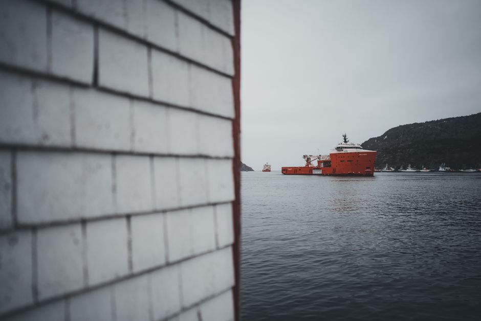 A close-up view of a white-painted brick wall with a textured surface occupies the left side of the image, showing horizontal rows of bricks with slightly weathered and uneven surfaces. The wall runs vertically alongside a narrow gap or corner. Behind the wall, a body of water extends into the distance, with a large, bright red offshore vessel floating prominently in the middle ground. The vessel has a smooth, metallic finish with visible structural details, including a bridge and superstructure at the aft. In the background, there are hilly landforms with sparse vegetation, and the sky is overcast with a greyish tone, creating a subdued, muted atmosphere. The scene suggests a waterfront or harbour setting, aligning with the context of private or non-local authority waste removal operations near a maritime environment, consistent with the themes of rubbish removal services for residential or commercial waterfront properties.