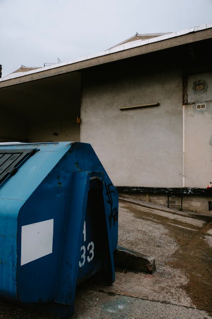 A large blue communal waste bin with a curved, textured metal surface showing signs of rust and weathering, positioned on a concrete pavement close to a building wall. The bin has a white rectangular label on its side and a numbered code partially visible, with black graffiti markings on the upper part. The ground around the bin appears damp and slightly stained. Behind it, the building's exterior wall is plain and off-white, with a black metal railing running along the base. The upper part of the building features a flat, overhanging roof with a minimalistic design, and a simplistic vertical pipe or support bracket is mounted on the wall. The environment is outdoors, under natural lighting with an overcast sky, and the scene aligns with on-site waste containment for private rubbish collection services, as managed by Waste Removal St John's Wood.