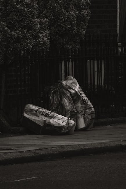 Three large black plastic garbage bags, absorbed in the dark and slightly reflective, are placed on a pavement near the curb in front of a black metal fence with vertical bars. The bags appear to be filled with waste materials, with some bulging and irregular shapes, indicating they are tightly packed. Behind the fence, dense foliage from leafy shrubs or trees is visible, adding a natural backdrop to the scene. The pavement shows signs of regular wear, with a concrete curb separating it from the road surface, which has a visible line marking. The lighting suggests an overcast or dimly lit environment, emphasizing the dark tones of the bags and surroundings. This image is typical of a private rubbish collection scene, possibly indicating an on-site clearance or independent waste disposal, often handled by waste removal services such as Waste Removal St John's Wood, to manage household or commercial waste outside of municipal collection schedules.
